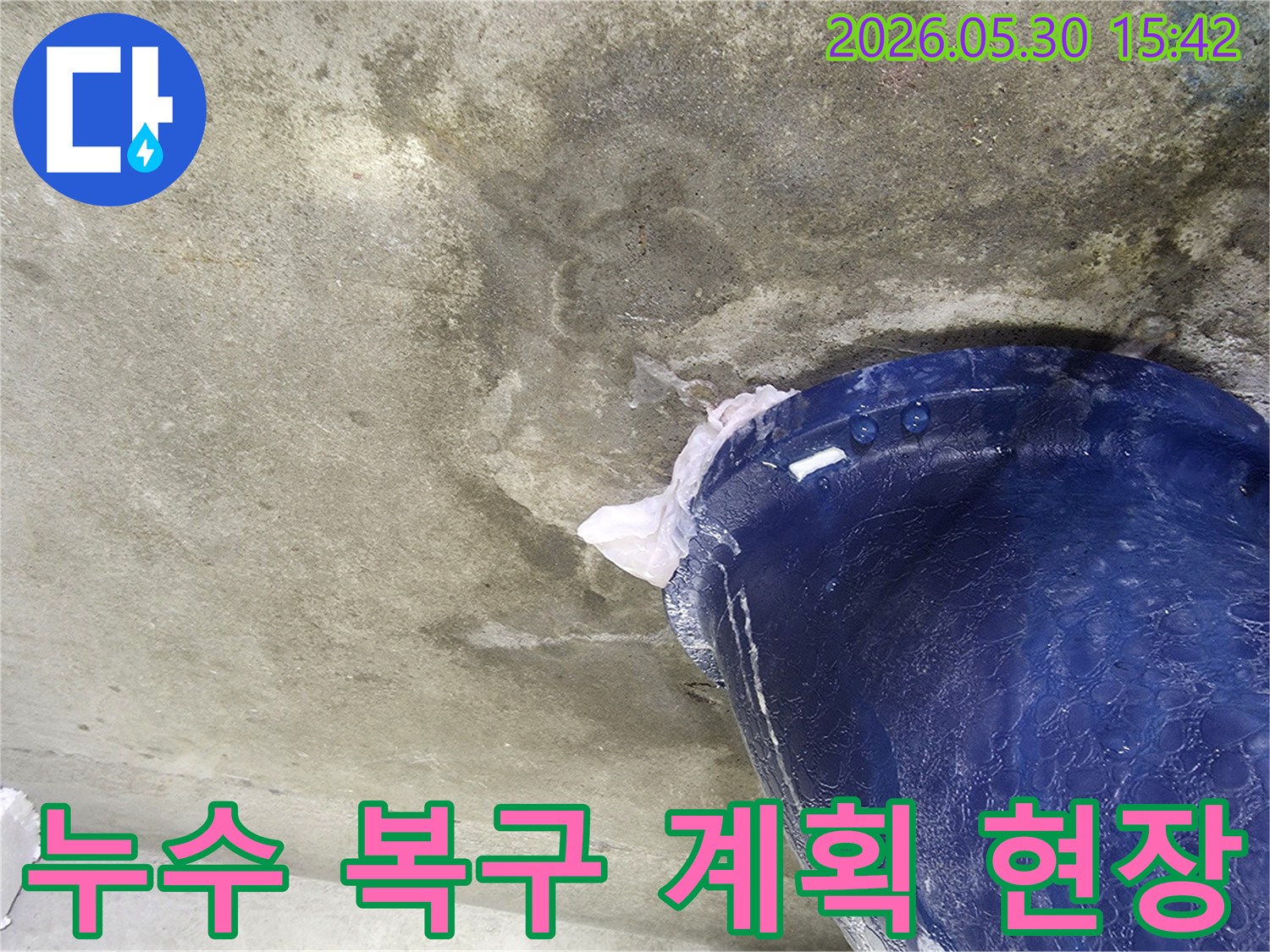 천장에서 물이 떨어지는 누수 현장에서-복구 계획 수립 전-추가 피해를 막기 위해-파란색 양동이로 물을 받아내는 모습입니다.