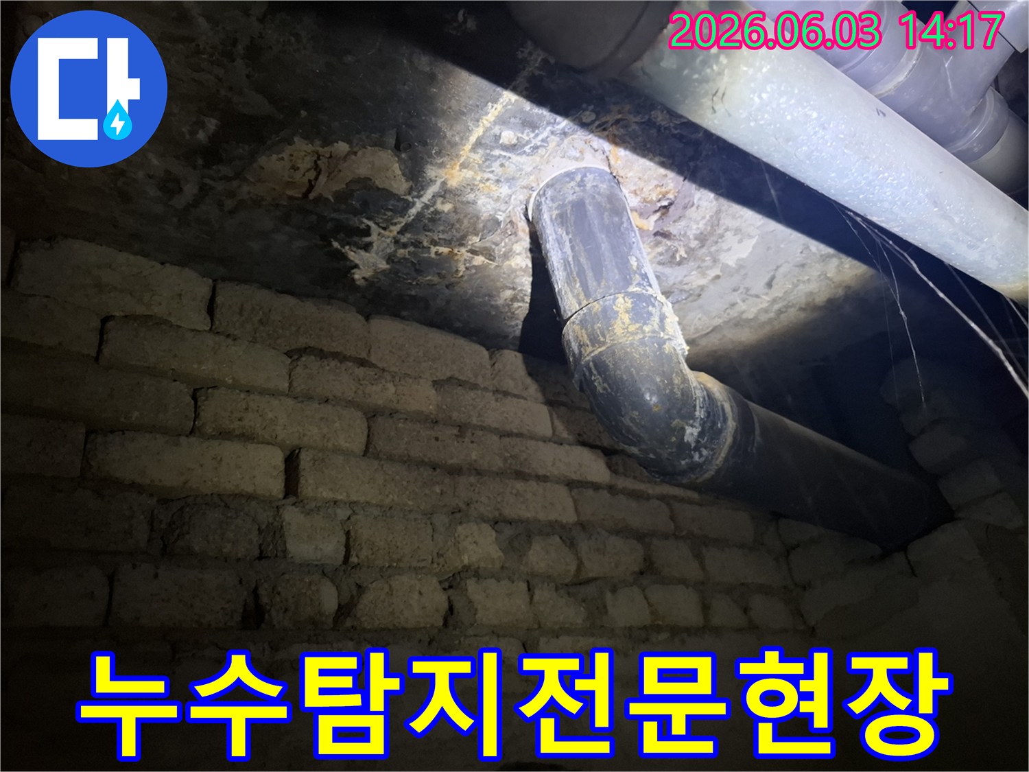 지하의 낡은 배관에서-물이 새어 나와-벽돌 벽에 스며든-누수 자국을-조명으로 비추어-면밀히 검사하는 모습