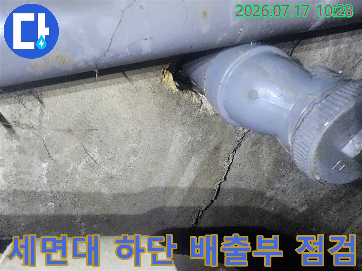 세면대 아래쪽 노출된 회색 배수 트랩과 주변 바닥의 균열 현황