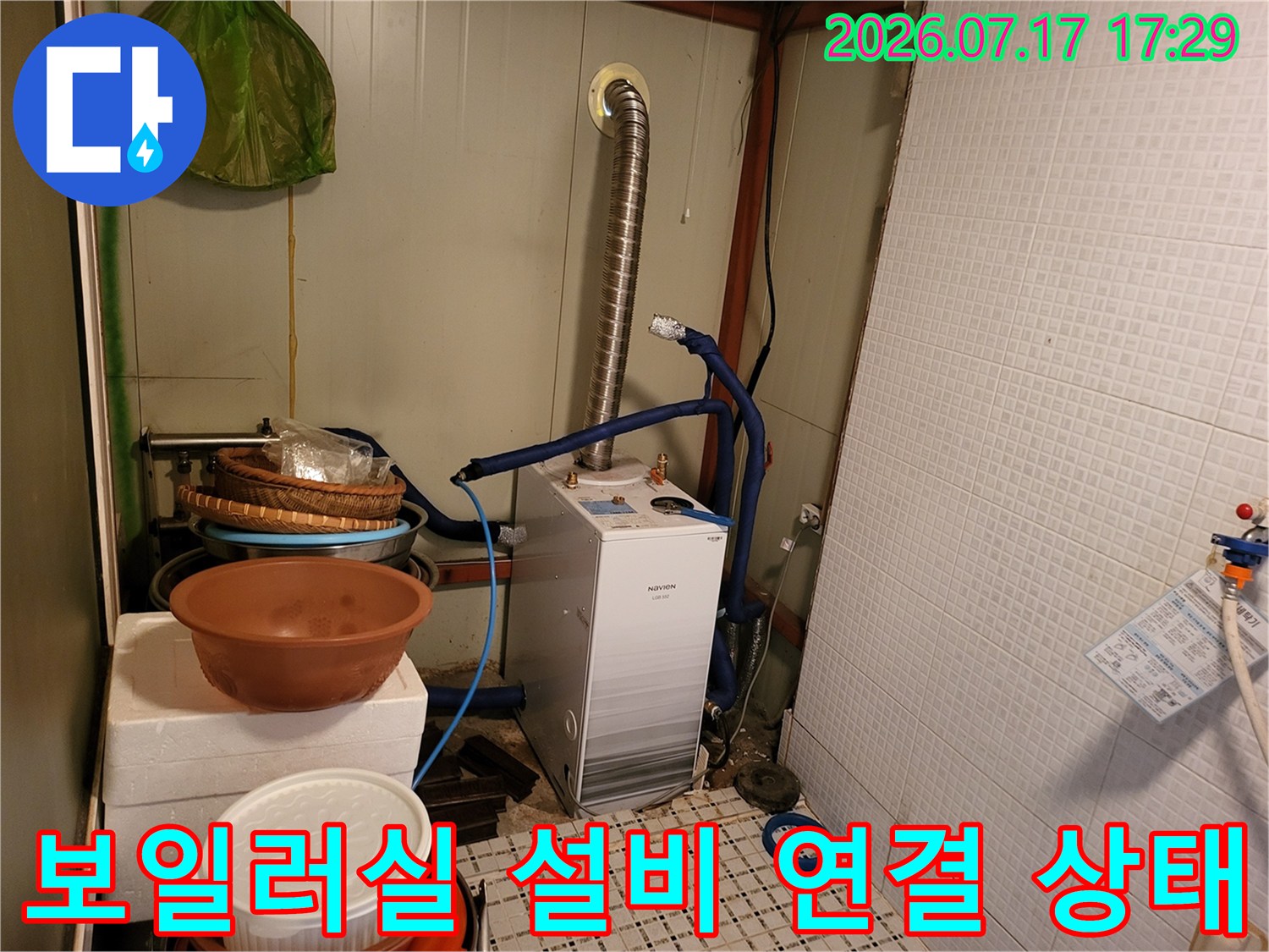 벽면 타일 앞의 흰색 보일러-상단 파란색 호스와 은색 자바라 관 연결 모습