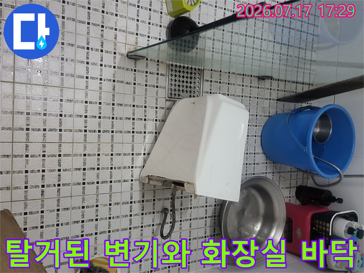 흰색 도기 변기가 타일 바닥에 놓여 있고 옆으로 급수 호스가 연결된-사진