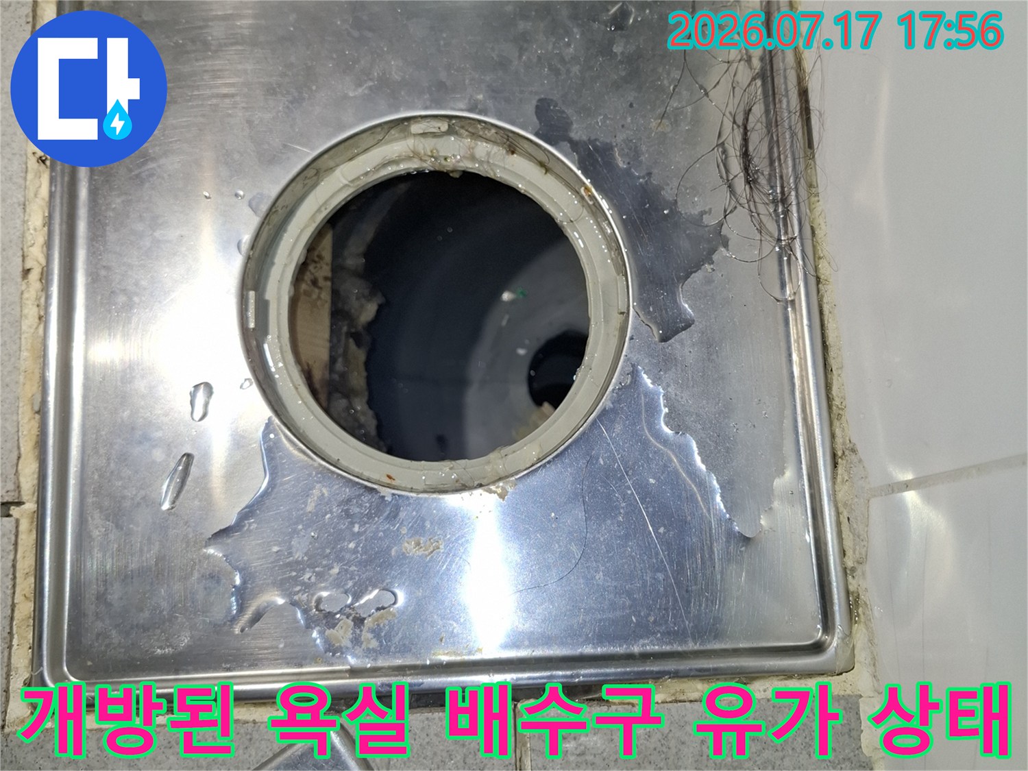 금속제 배수구 덮개가 옆으로 치워져 있고-어두운 배관 내부가 보이는 화장실 바닥 사진