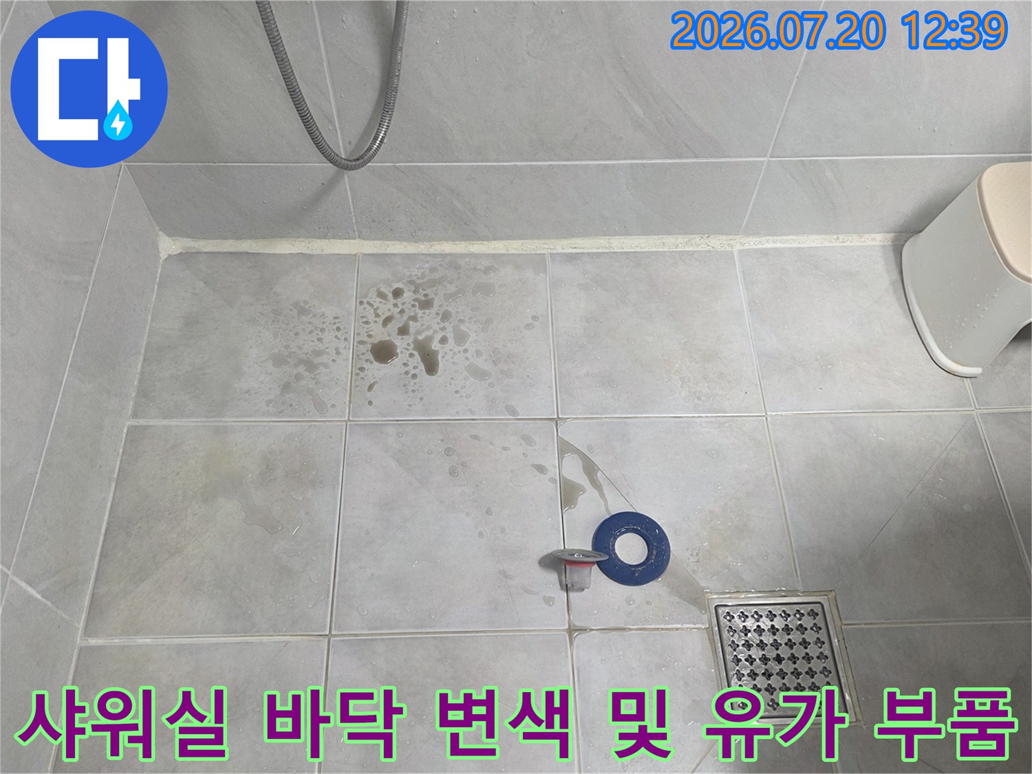흰색 욕실 타일 위에 갈색의 습기 얼룩이 있고 하수구 구멍 옆에 파란 링이 있는 모습