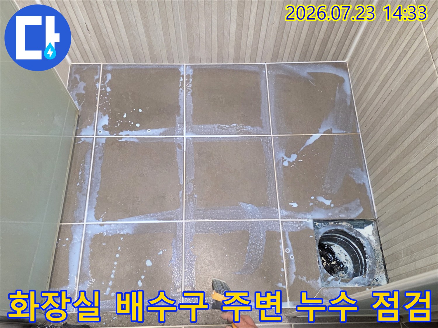 욕실 바닥 타일 위로 하얀색 세제 거품이 퍼져 있고-우측 하단에 검은색 배수구 유가가 보이는 모습
