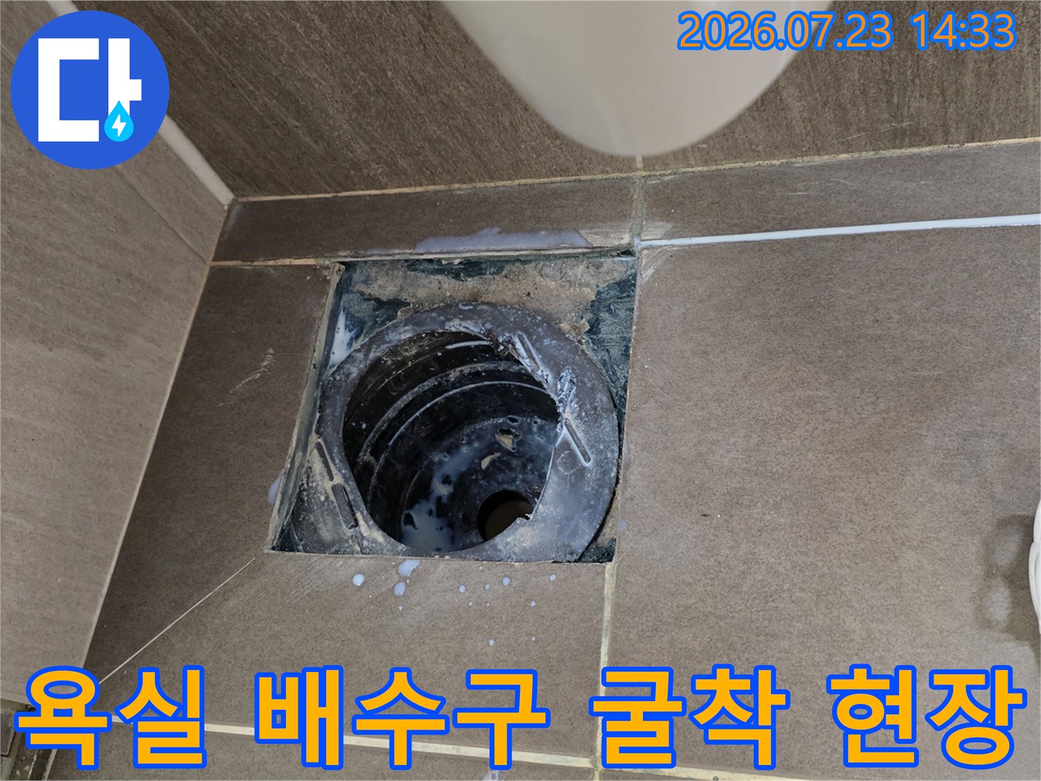 화장실 바닥 타일을 일부 제거하고 내부의 검은색 원형 배수관이 드러난 모습