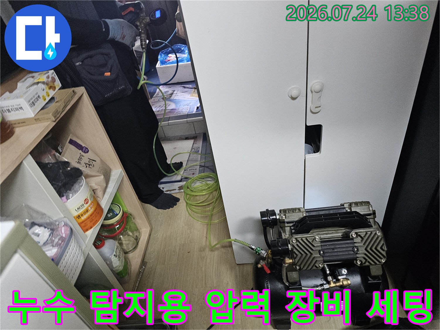 흰색 수납장 앞에 놓인 검은색 누수 탐지용 콤프레셔와 연결된 초록색 호스