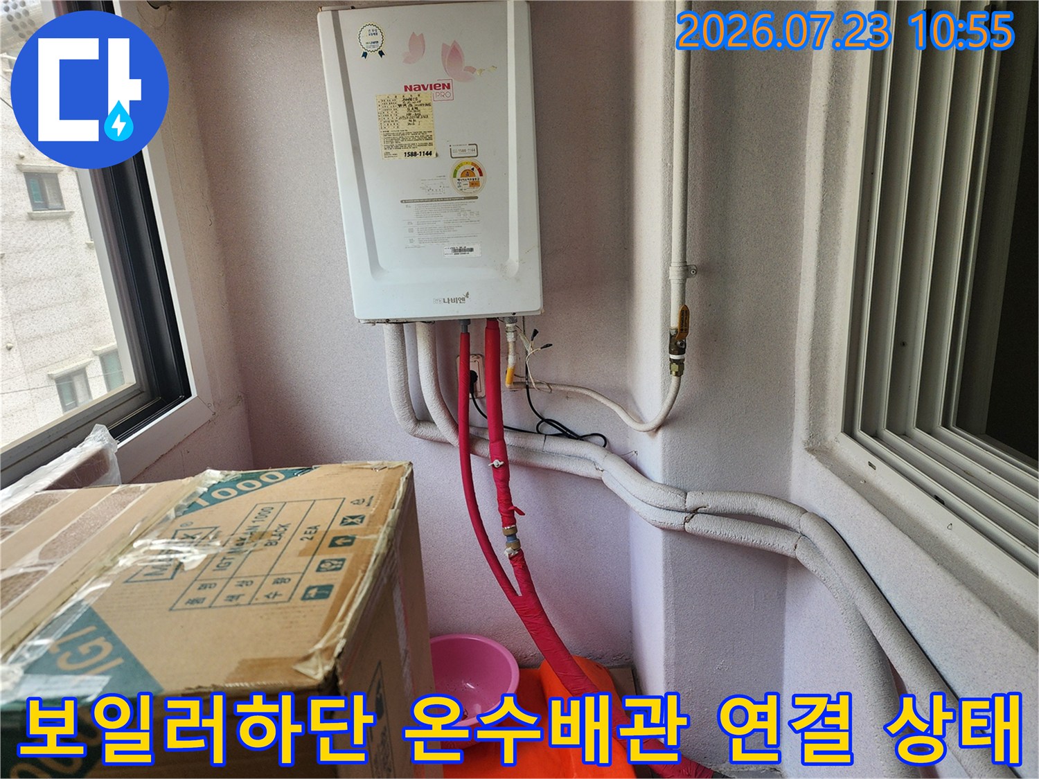 흰색 벽면에 설치된 가스 보일러와 그 아래로 연결된 붉은색 온수 배관-회색 보온재 배관의 모습
