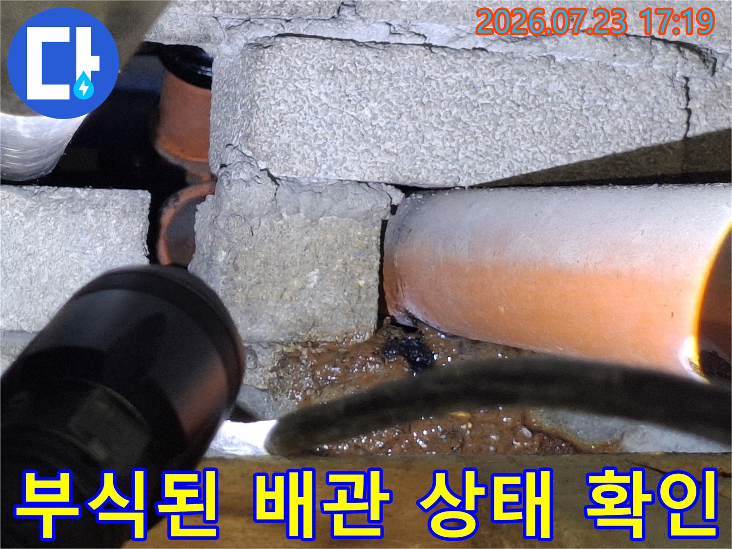 콘크리트 구조물 아래에 위치한 오렌지색 배관의 부식된 외벽 모습