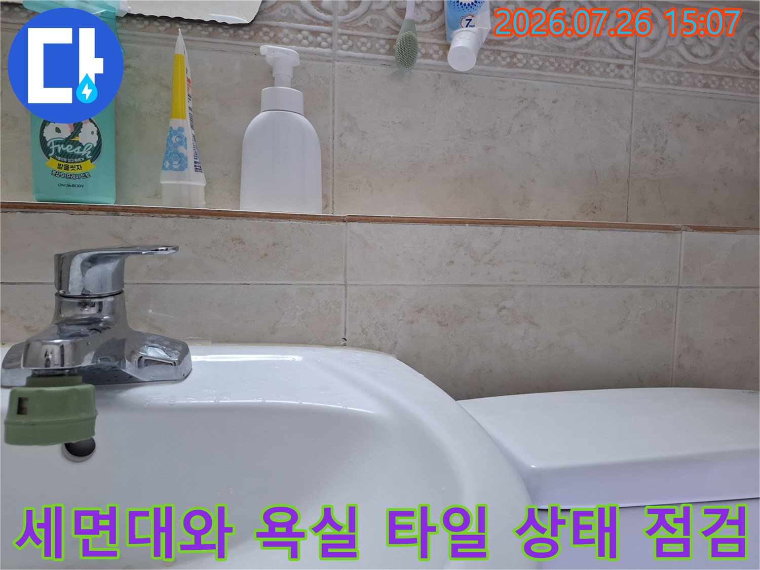 흰색 세면대 수전과 그 뒤로 보이는 베이지색 욕실 타일-벽면 선반 위에 놓인 세정제와 칫솔들