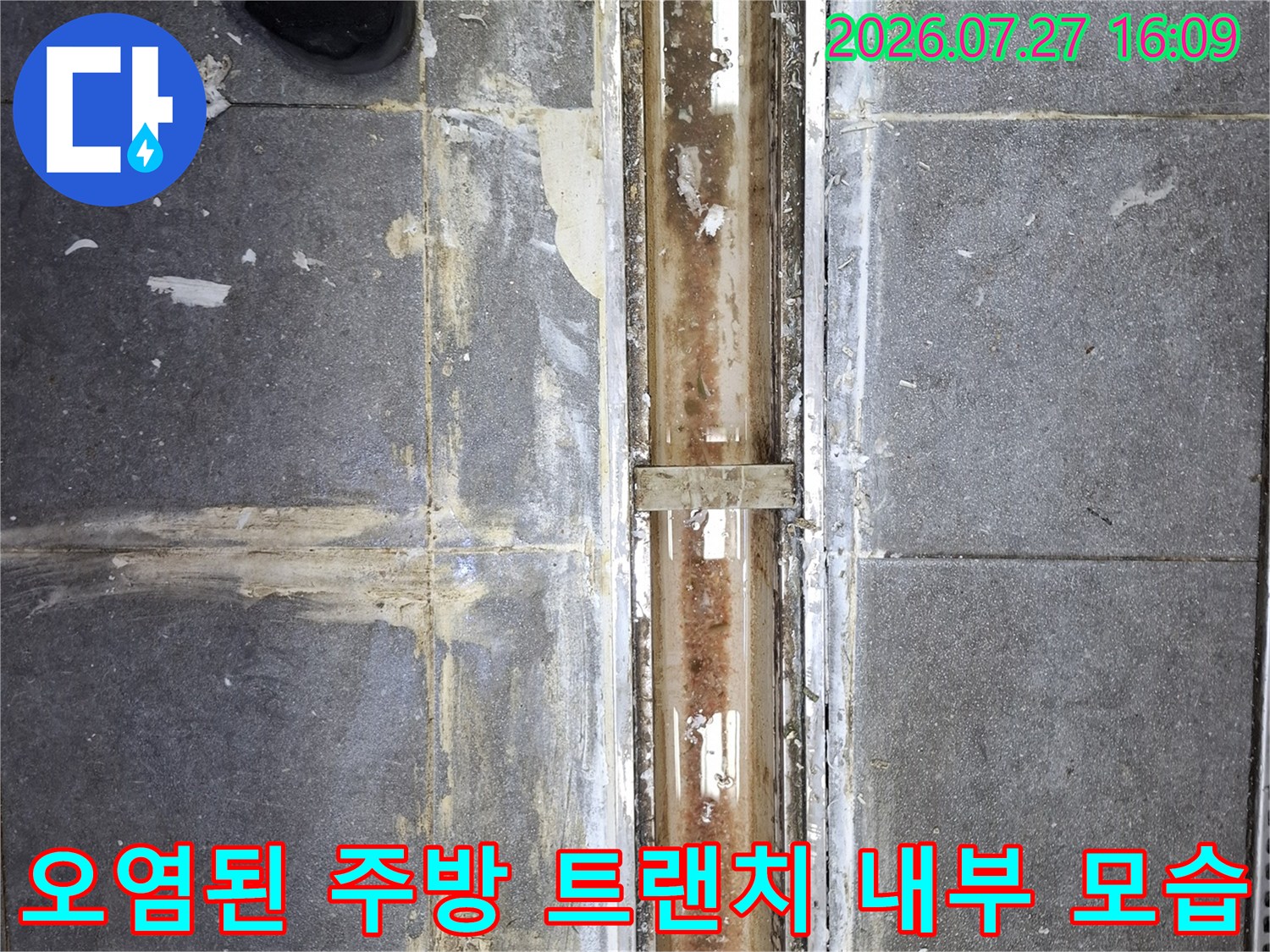 금속제 배수관 구조물이 보이며 주변에 하얀색 백화 현상과 오염이 있는 주방 바닥 사진
