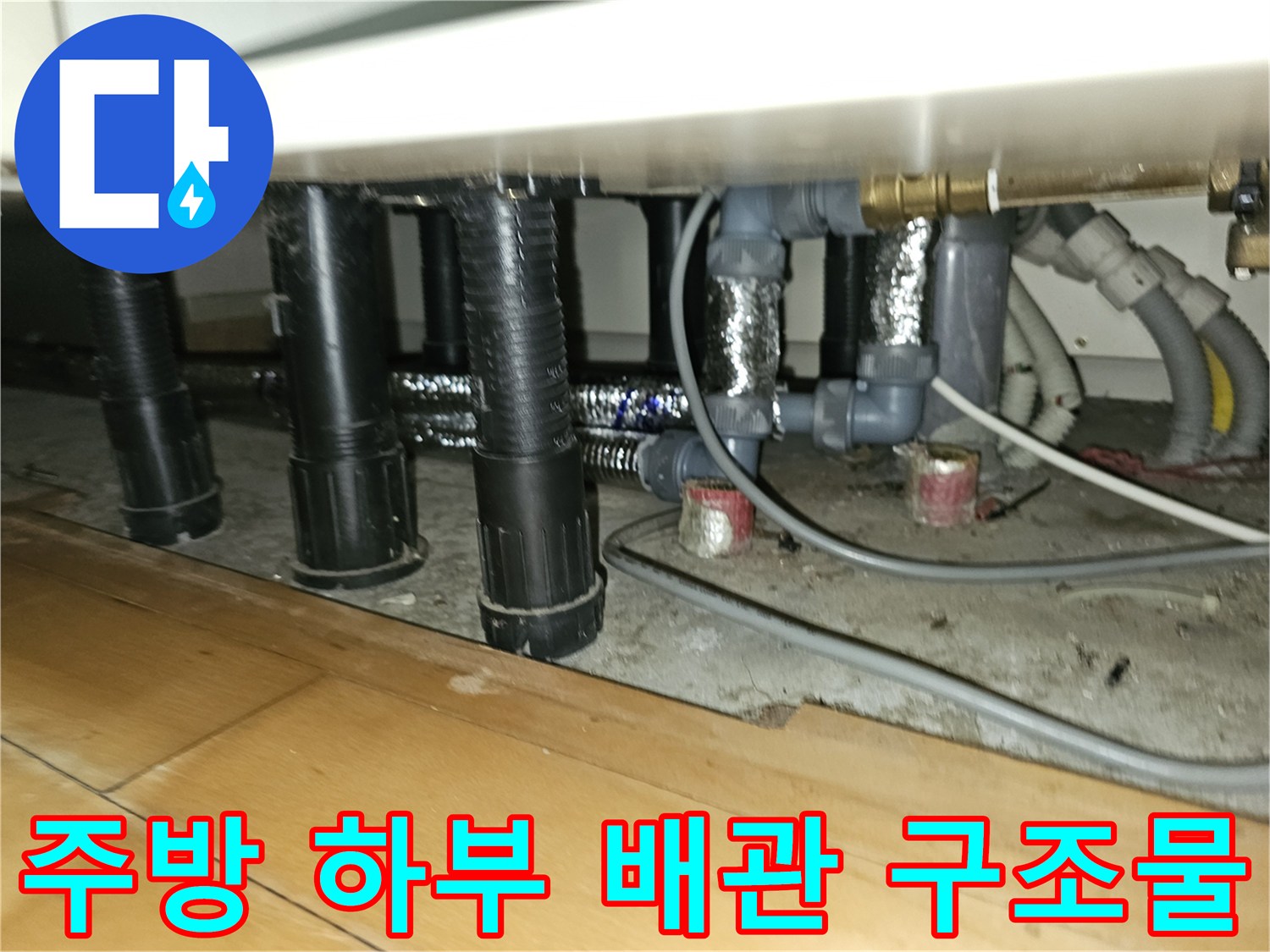검은색 플라스틱 관과 은색 보온재가 섞인 주방 하부장 배관 모습입니다.