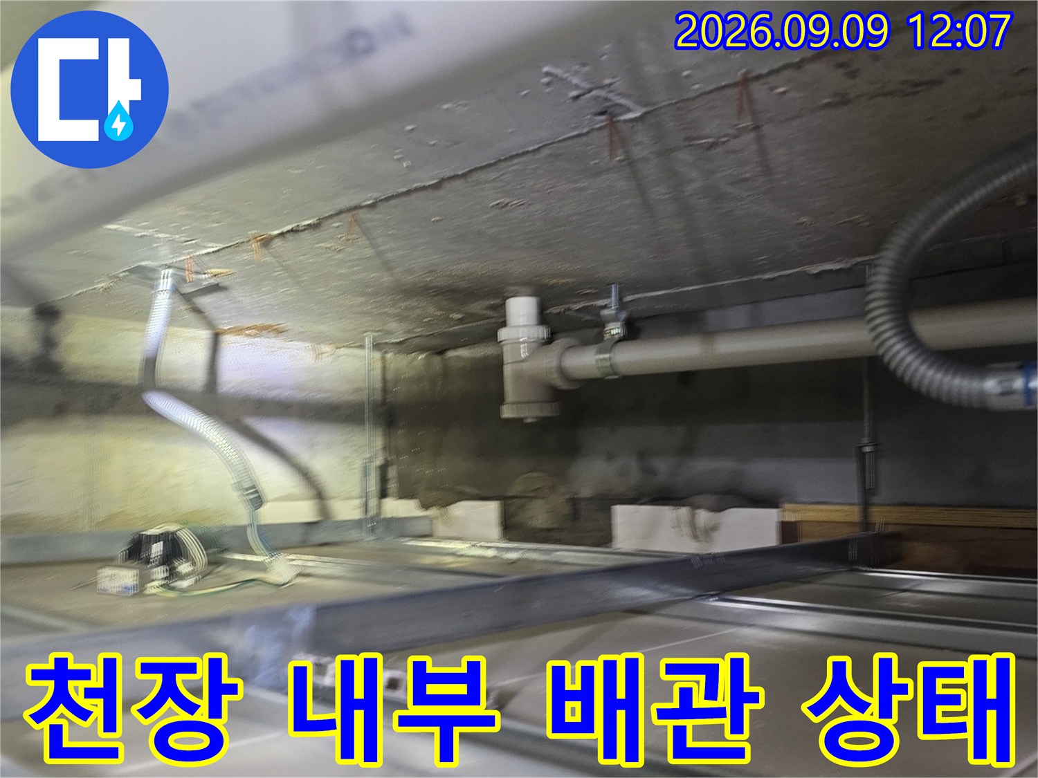 천장 마감재 안쪽의 굵은 배관과 은색 주름관이 노출된 현장 사진입니다.